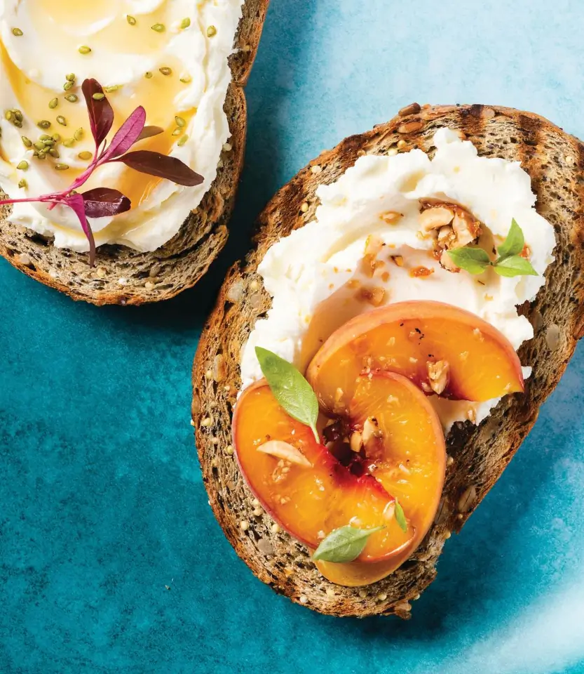 Two slices of seeded whole grain toast topped with cream cheese, fresh peaches, and garnished with nuts and microgreens on a turquoise surface.