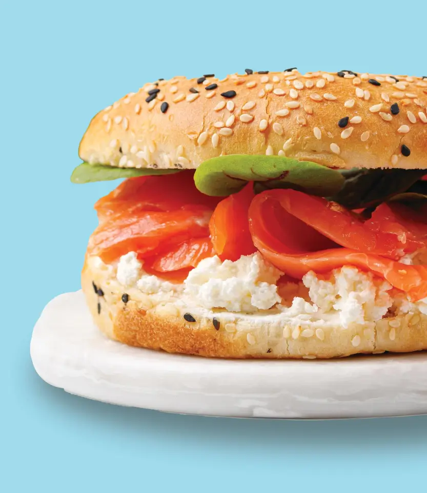 Bagel sandwich with cream cheese, smoked salmon, and avocado slices on a white plate against a blue background.