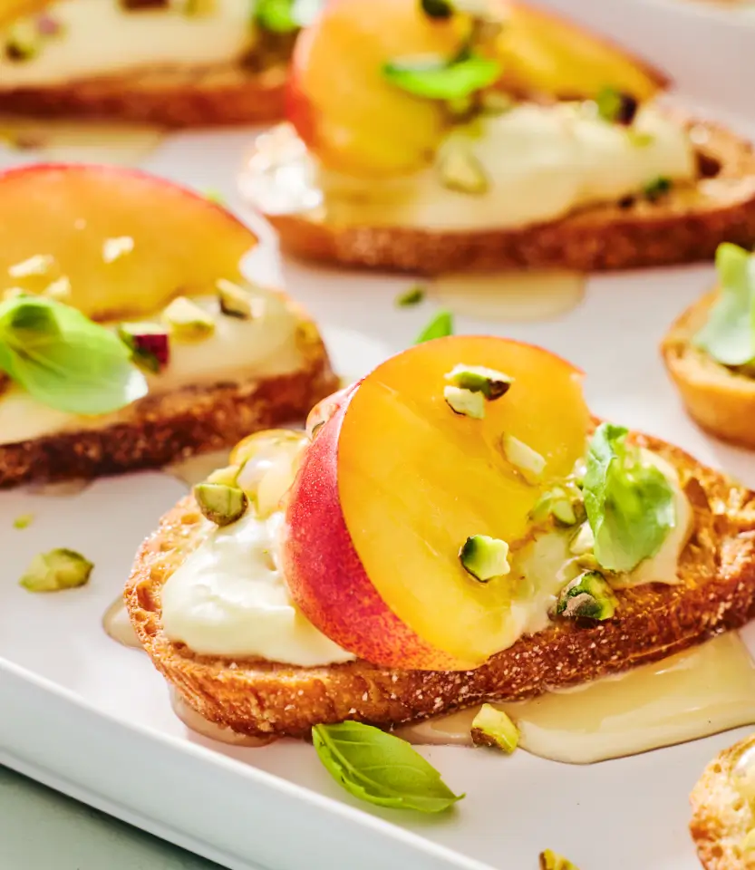 Gourmet open-faced sandwiches on toasted bread with cream cheese, yellow and red stone fruit slices, and pistachio garnish on a white plate.