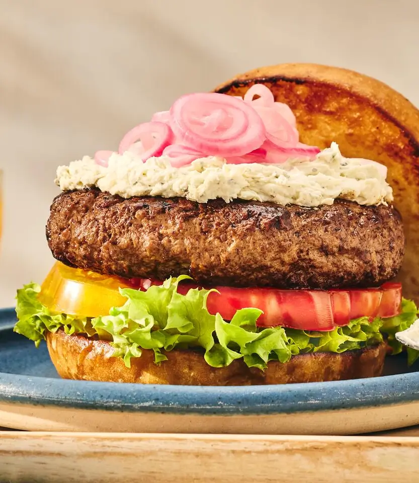 Gourmet burger with beef patty, lettuce, tomato, yellow pepper, white mayo, pink pickled onions, and toasted bun on blue plate.