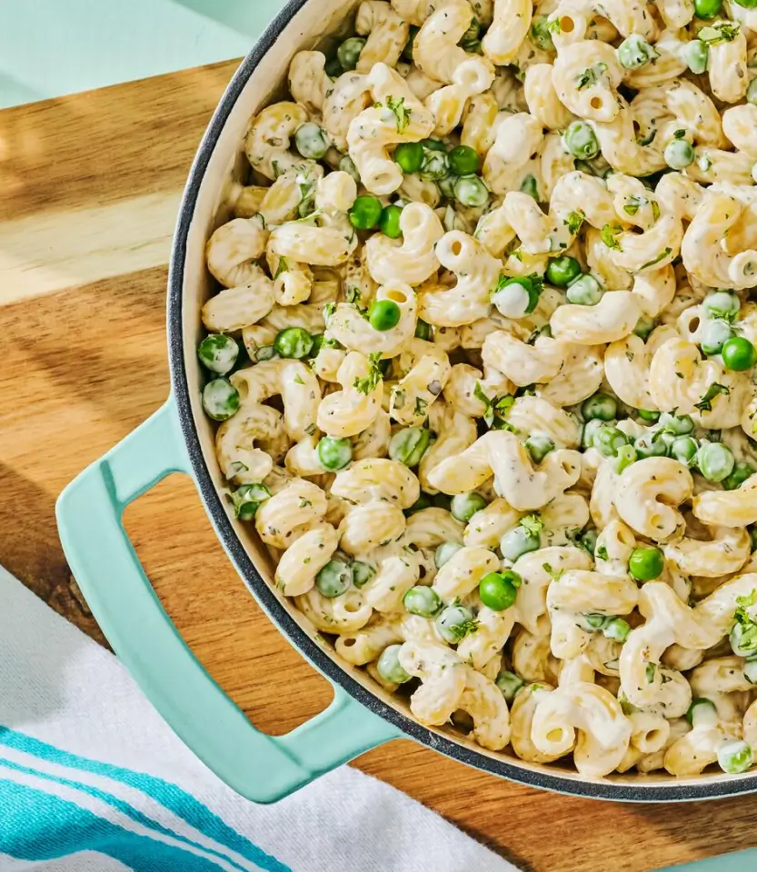 Creamy pasta salad with elbow macaroni, peas, and fresh herbs in a turquoise Dutch oven on a wooden board.