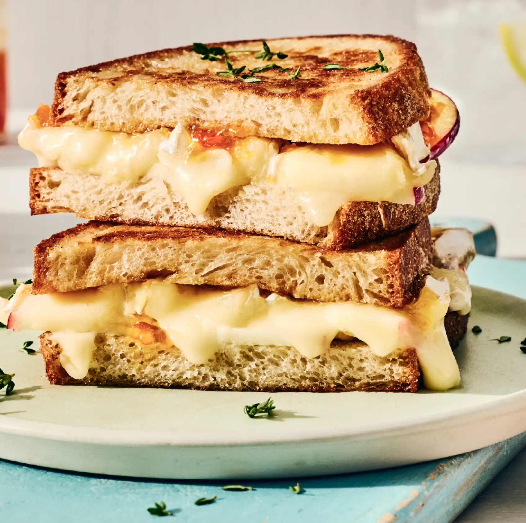 Stacked grilled cheese sandwich with melted cheese and sliced apple on toasted bread, garnished with fresh thyme on a white plate.