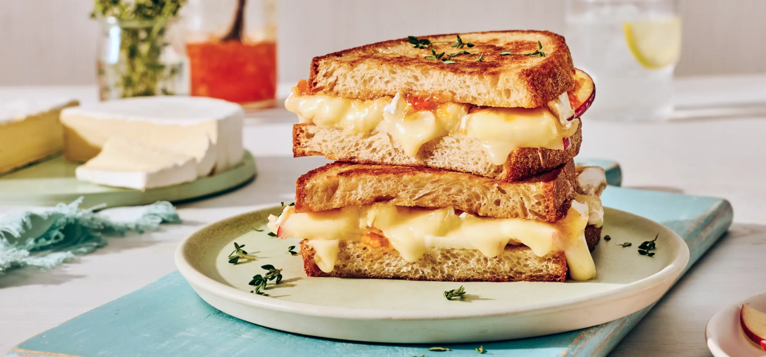 Stacked grilled cheese sandwich with melted cheese and red onions on a white plate, garnished with fresh thyme, surrounded by cheese wheels and beverages.