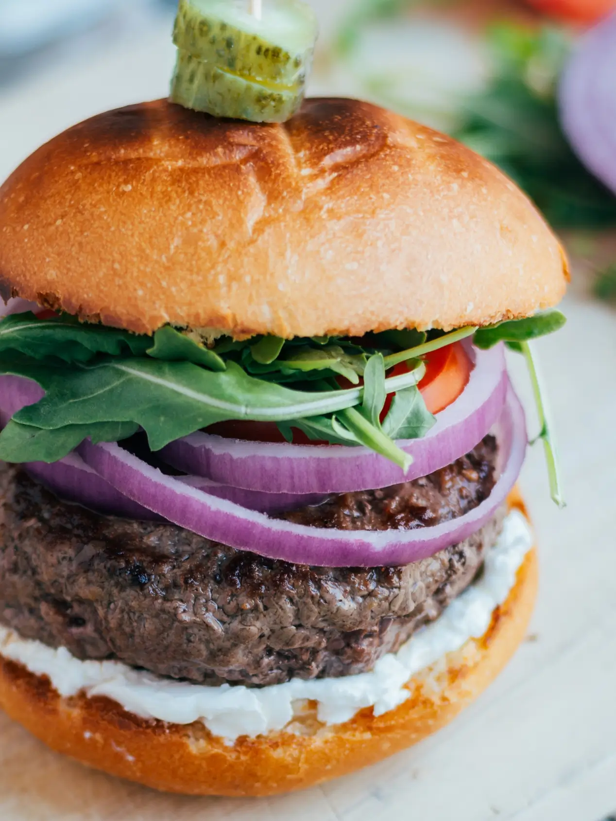 Gourmet burger with toasted bun, beef patty, white sauce, purple onions, arugula, tomato, and cucumber garnish on top.