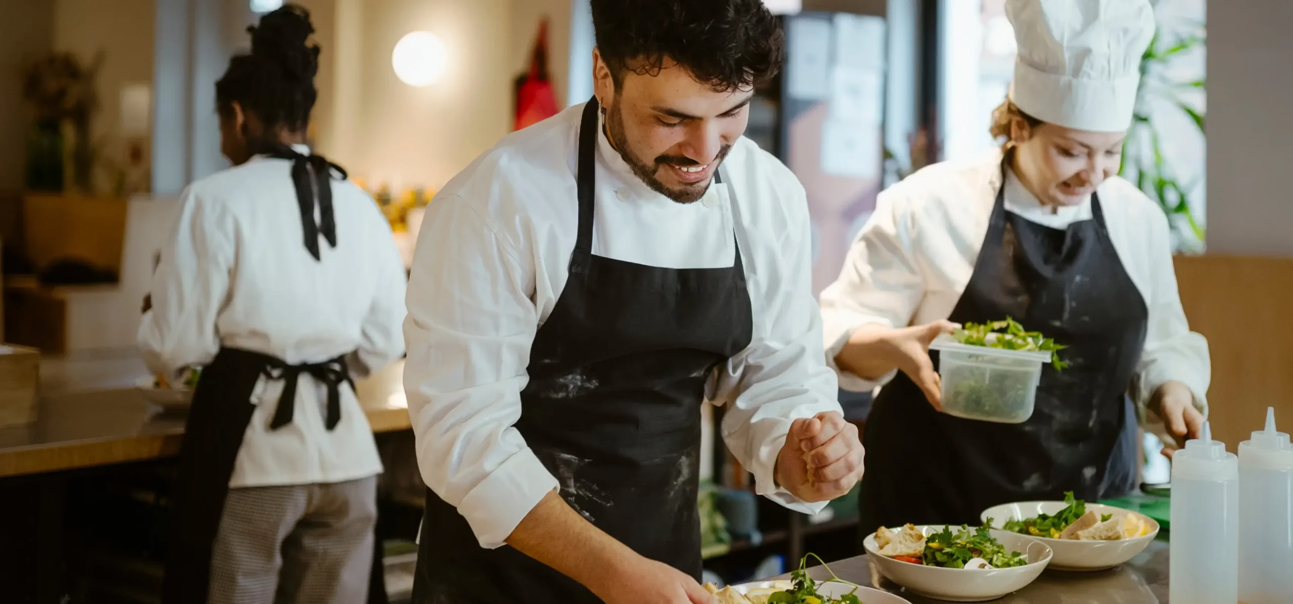 Professional chefs in white coats and black aprons prepare plated dishes in a kitchen, with fresh salads and garnishes visible on the counter.