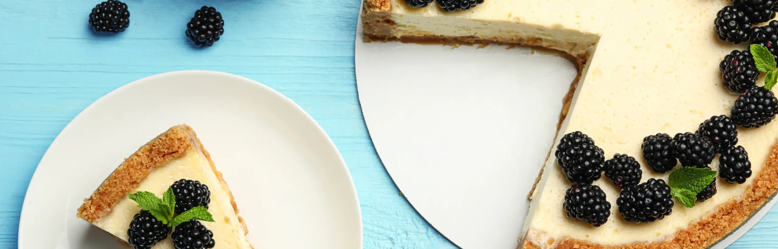 Cheesecake slices with blackberries and mint on a blue wooden surface, featuring a whole cake with fresh berries.