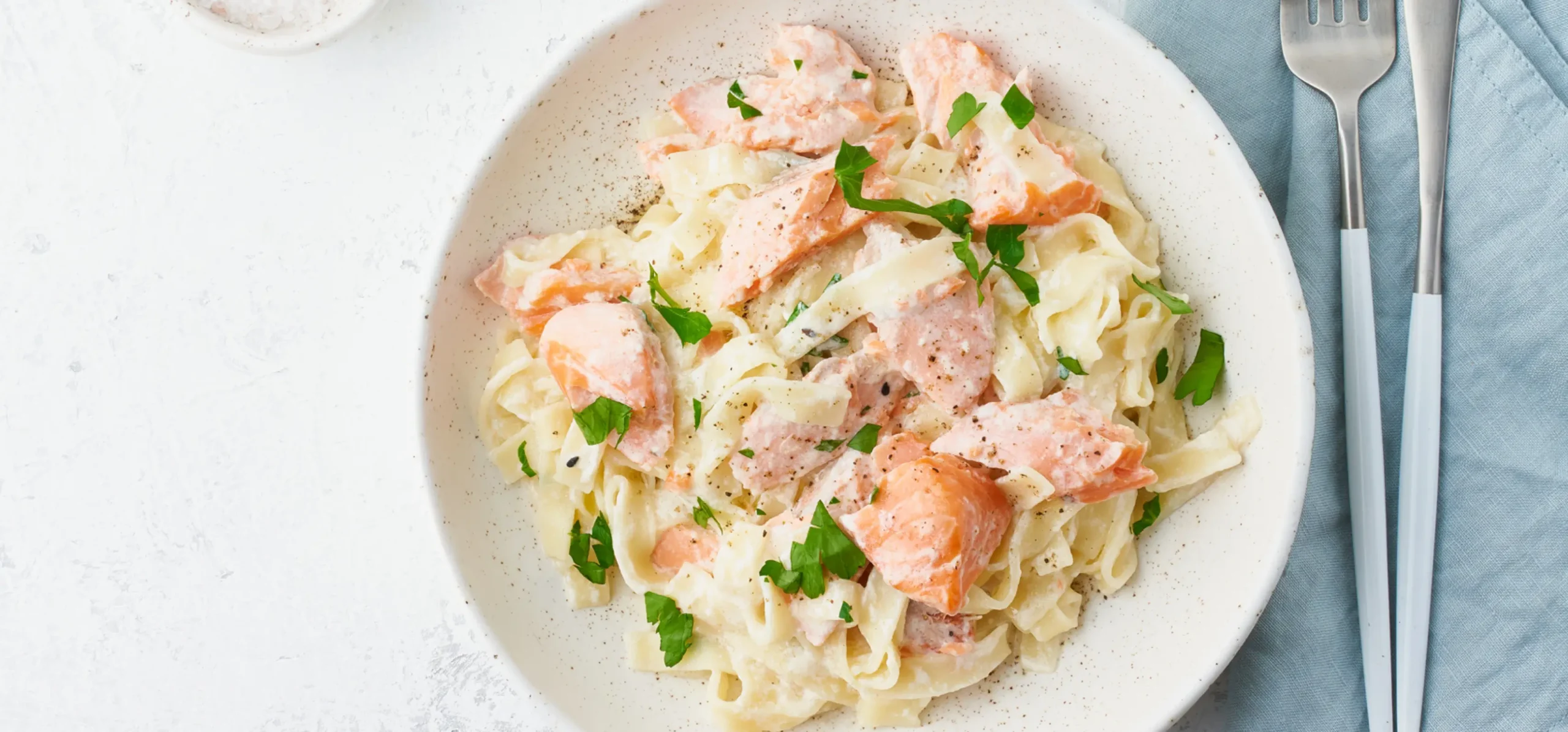 Pasta with smoked salmon, cream sauce, and fresh parsley garnish on a speckled white plate with fork and knife.