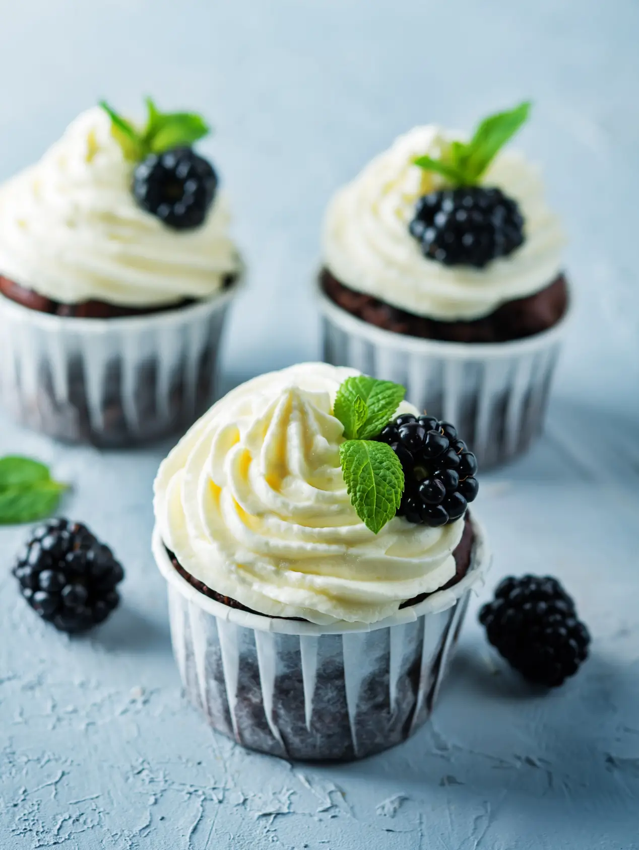 Three chocolate cupcakes with cream frosting, fresh blackberries, and mint leaves on a light blue textured surface.