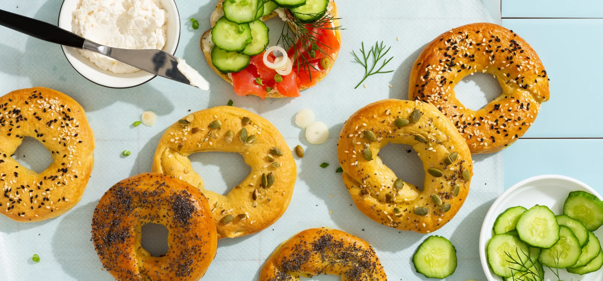 Assorted seeded bagels arranged with cream cheese, fresh cucumber slices, smoked salmon, and dill garnish on a light blue surface.