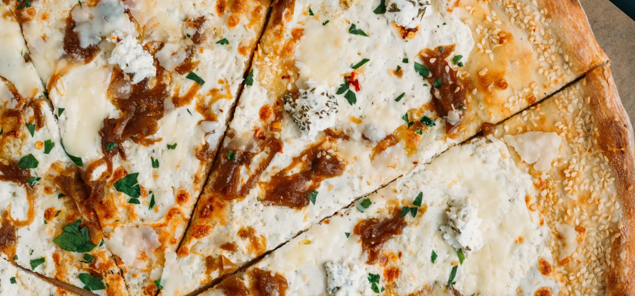 Sliced flatbread topped with melted cheese, caramelized onions, fresh herbs, and seasoning on a wooden surface.