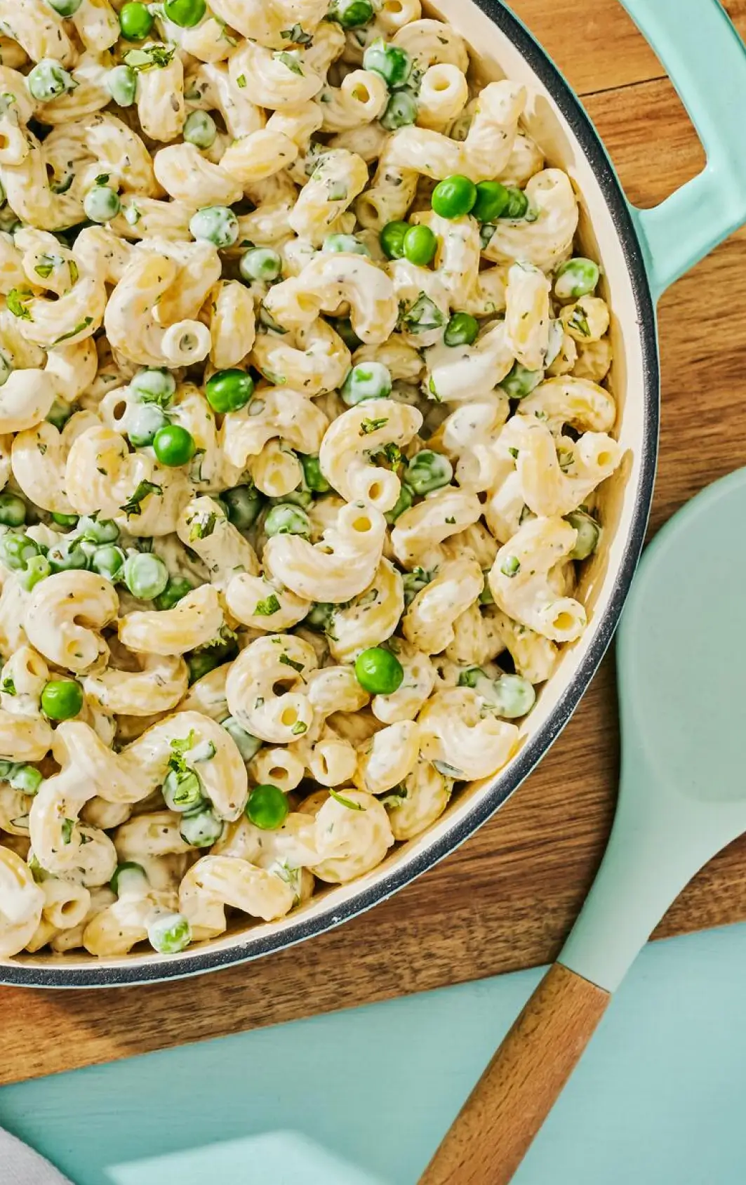 Creamy pasta salad with ring-shaped pasta, green peas, sliced green onions, and black pepper in a light blue skillet on a wooden board.