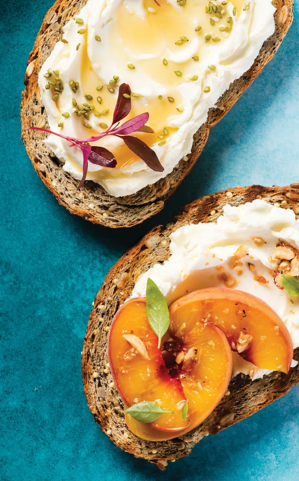 Two open-faced toasts on teal background: top slice with ricotta, honey, pumpkin seeds, and edible flowers; bottom slice with ricotta, sliced apricots, and nuts.