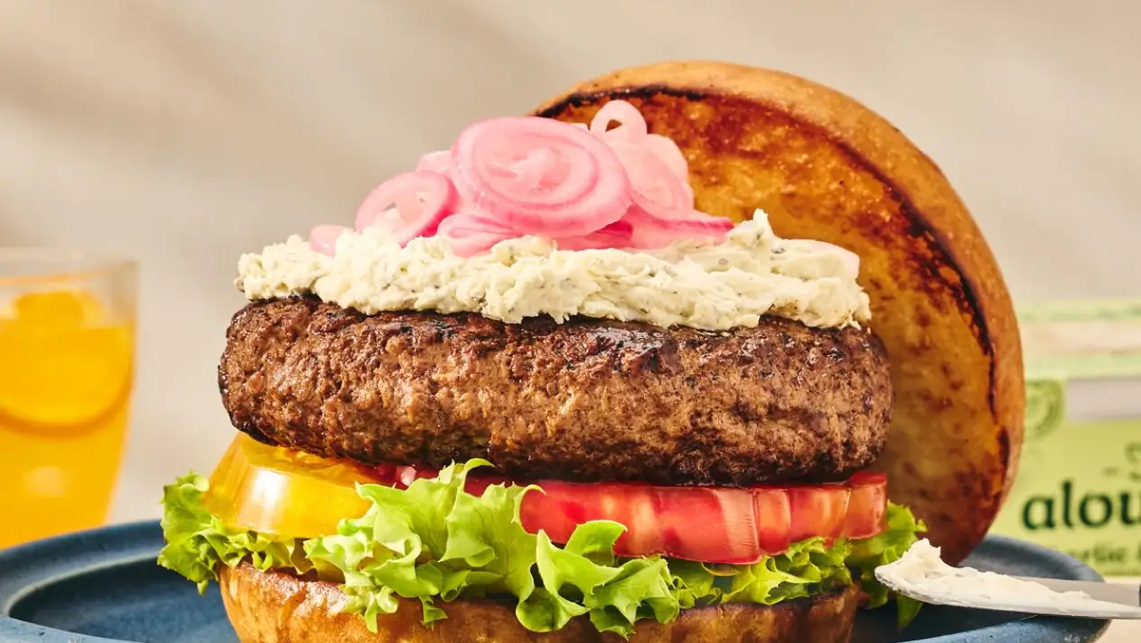 A gourmet burger on a toasted bun with lettuce, tomato, white sauce, and pickled pink onions, served on a blue plate with a beverage in the background.